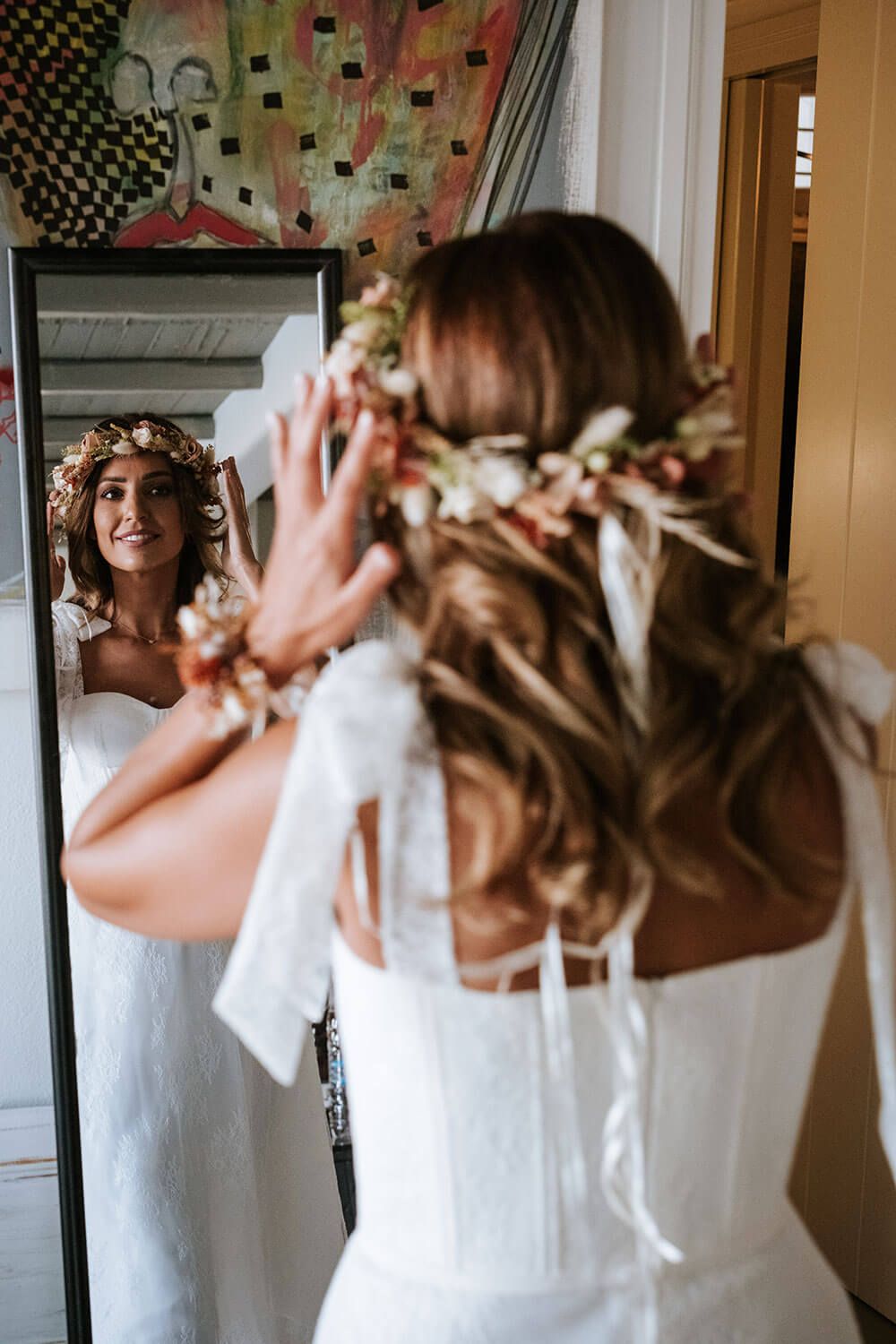 A bride in a white wedding dress looks at her reflection in a mirror while wearing a delicate floral crown.