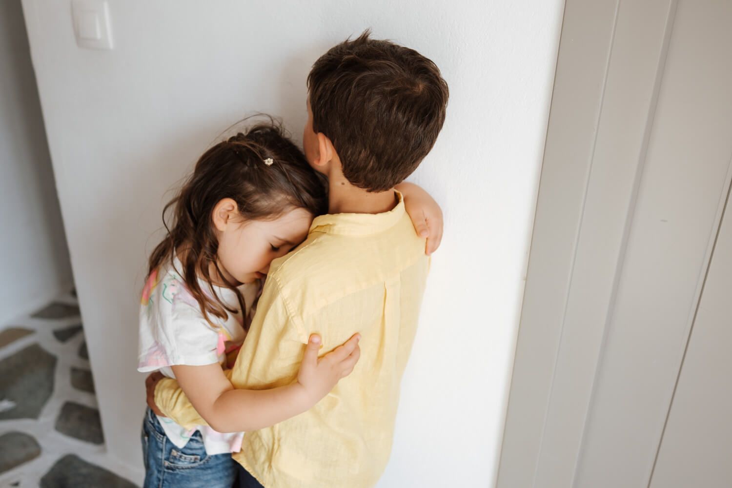 tender moments showing two kids embracing against a light-colored wall.