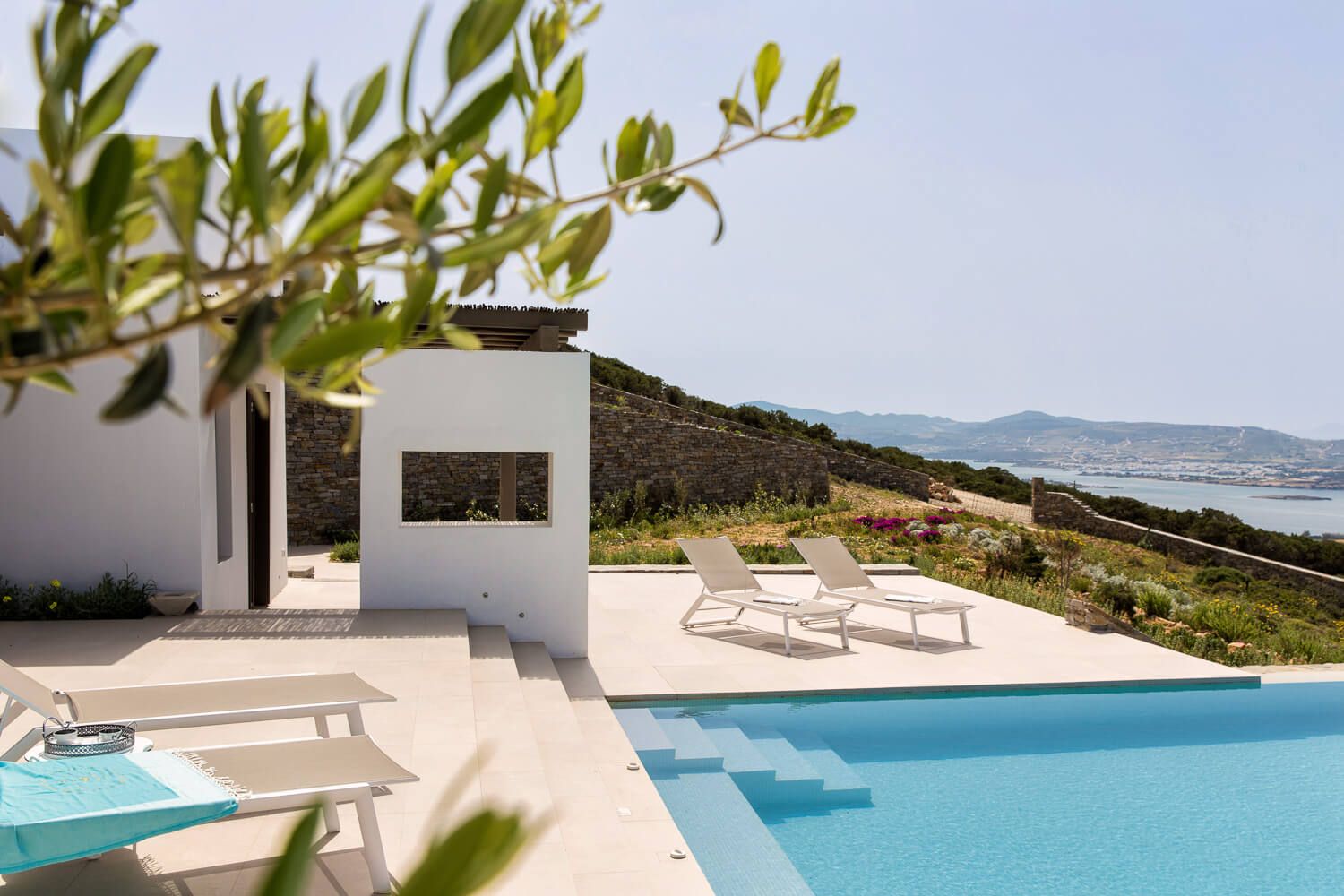 Luxury infinity pool overlooking a scenic Aegean coastline with white architecture and trees.