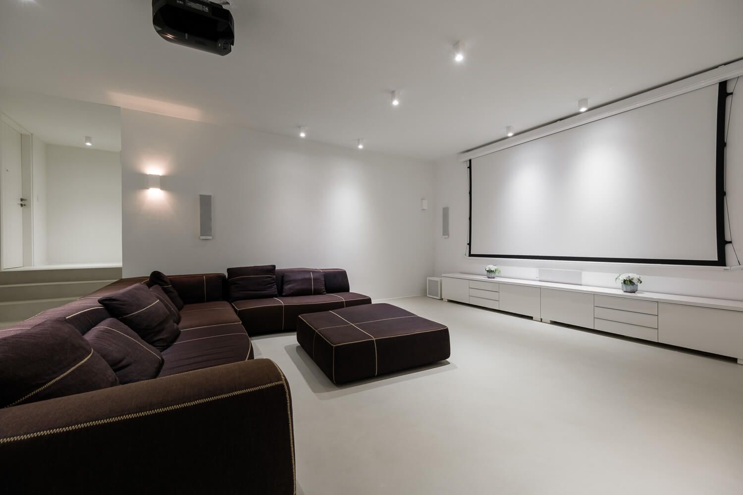 Modern home theater room with dark brown sectional sofa and white walls featuring ceiling mounted projector screen.
