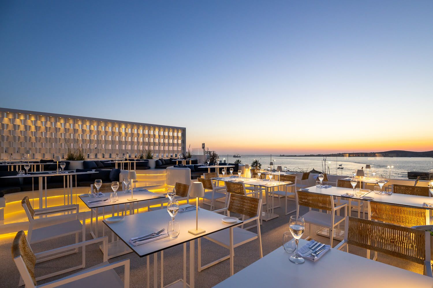 Elegant rooftop restaurant terrace overlooking a shimmering bay at golden sunset in Paros island.