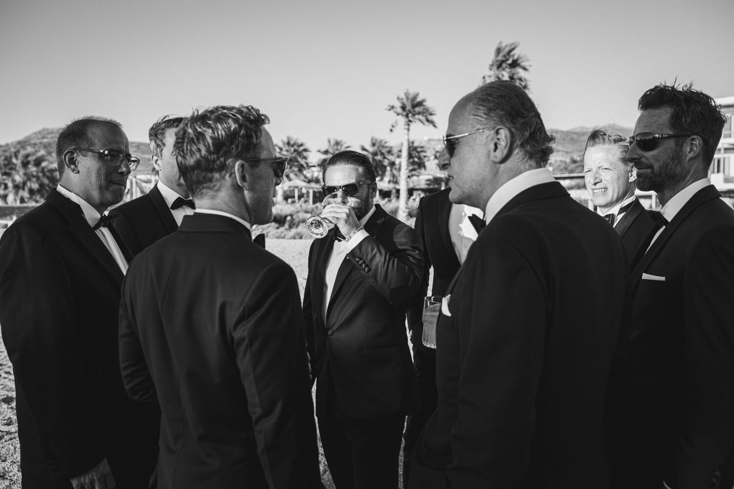 Black and white photo of groomsmen in suits conversing outdoors with palm trees in the background.