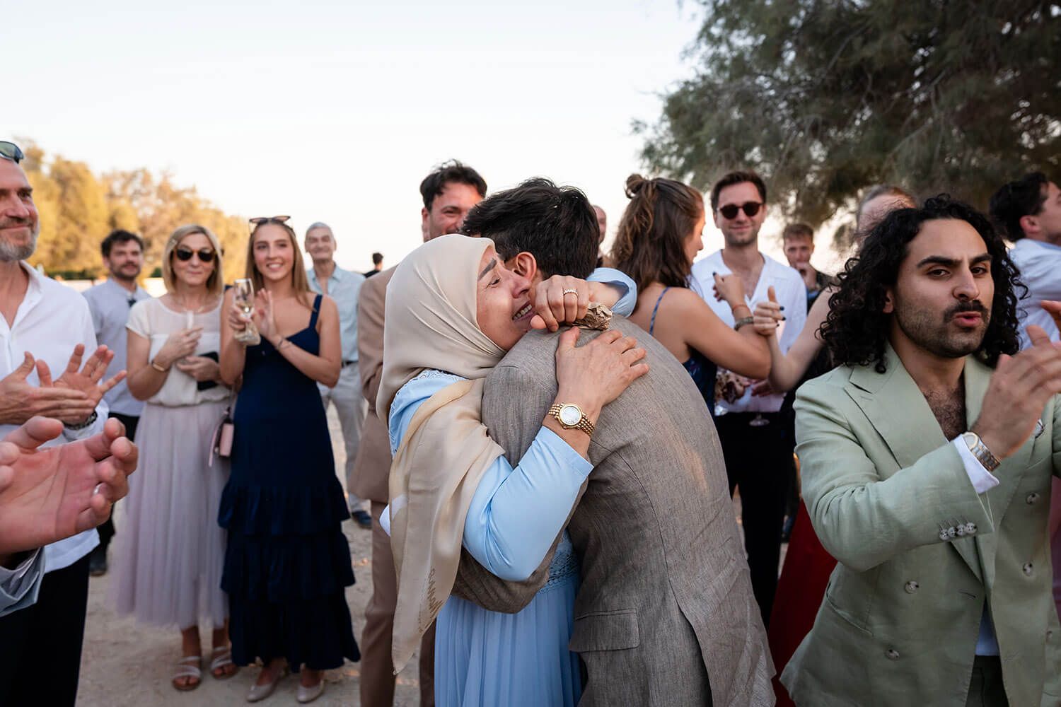 Wedding guests clap and celebrate during an outdoor sunset ceremony while thr groom embraces his mother in the center.