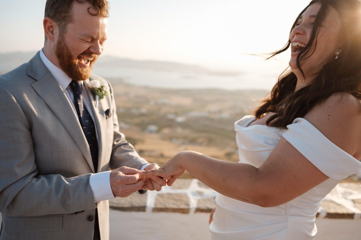 Groom placing ring on brides finger during outdoor ceremony with scenic sunset backdrop in Antiparos