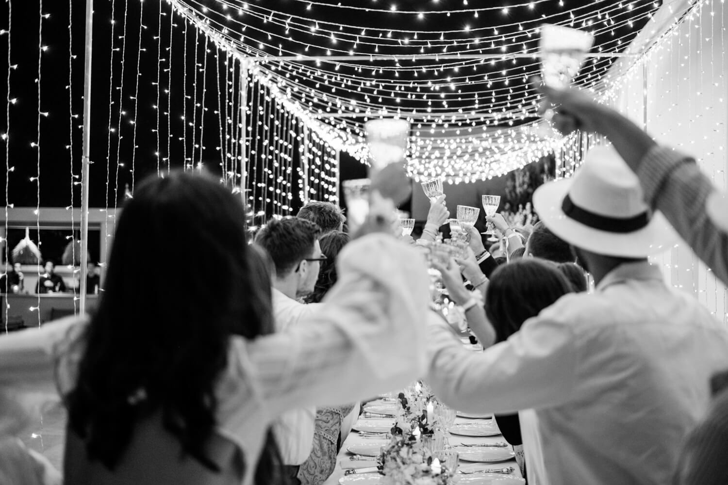 Black and white wedding dinner scene with guests raising hands under canopy of twinkling lights.