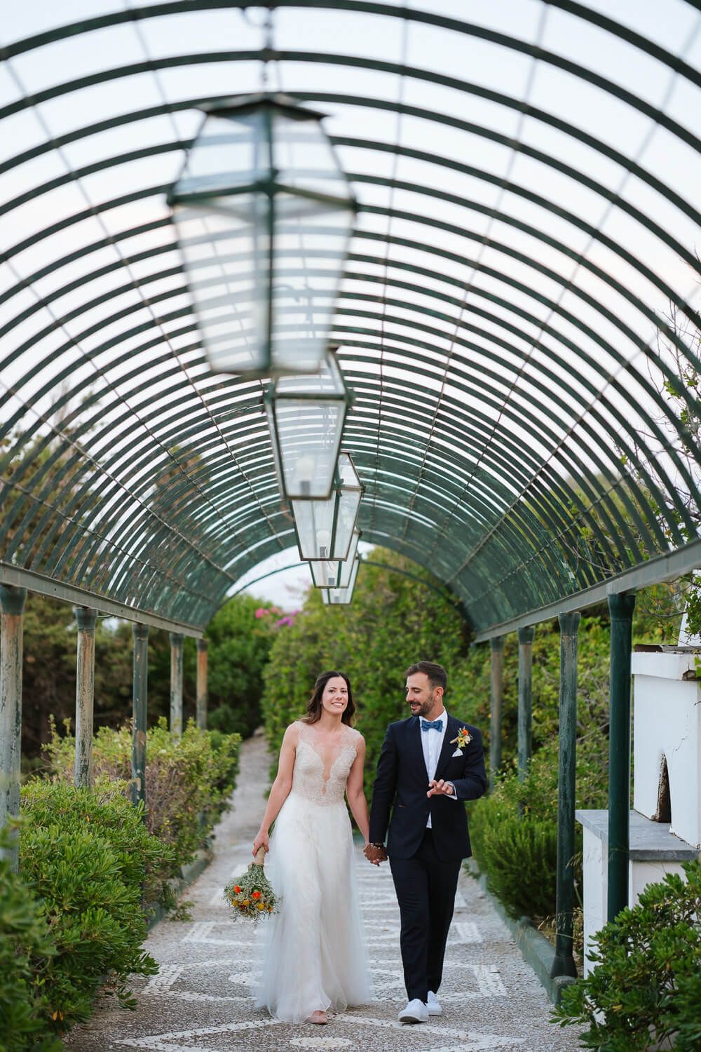 Bride and groom walk hand in hand through elegant arched garden tunnel in Syros Greece.