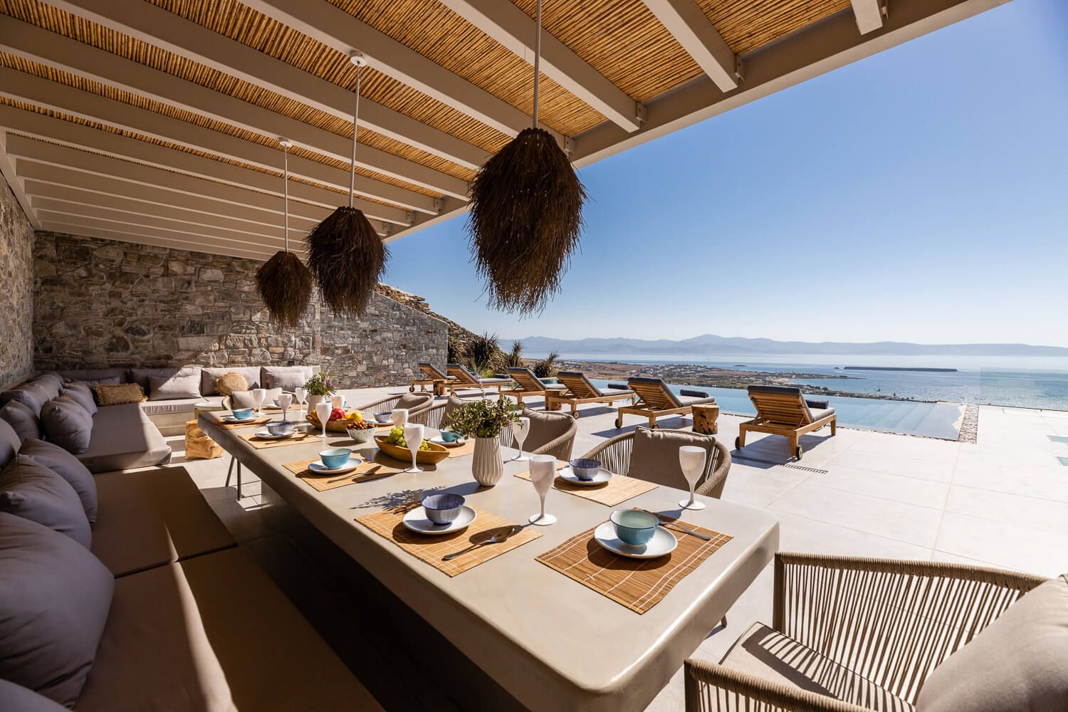 Outdoor dining area with long table, sea views, woven pendant lights, and stone wall in Paros island