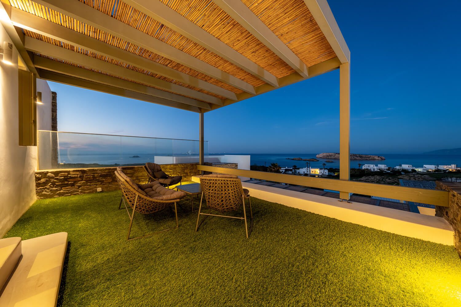Panoramic view of a modern covered patio with wooden ceiling and artificial grass overlooking the ocean at dusk.