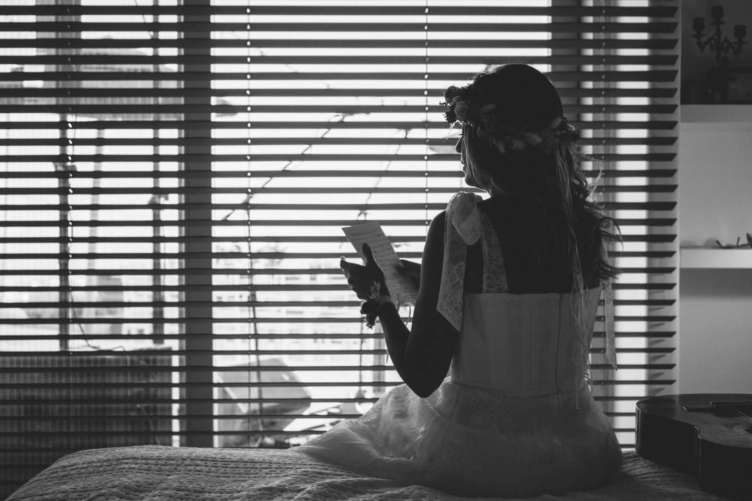 Bride in wedding dress sits by window reading in atmospheric black and white silhouette photo.