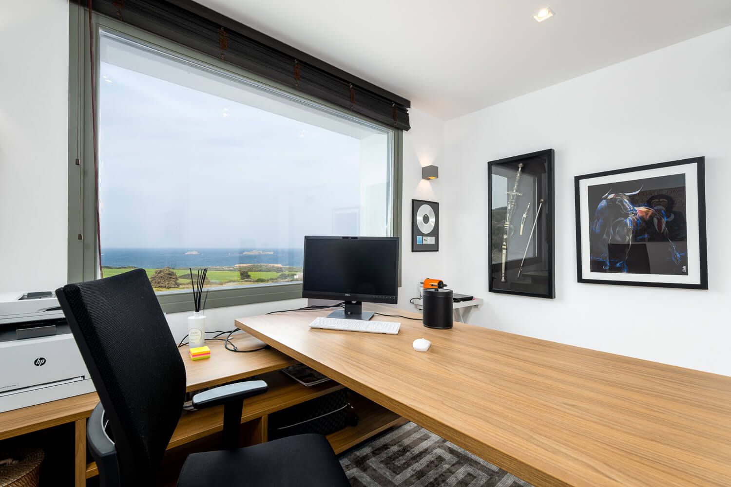 Modern home office setup with light wood desk, black chair, and window view of coastal landscape.