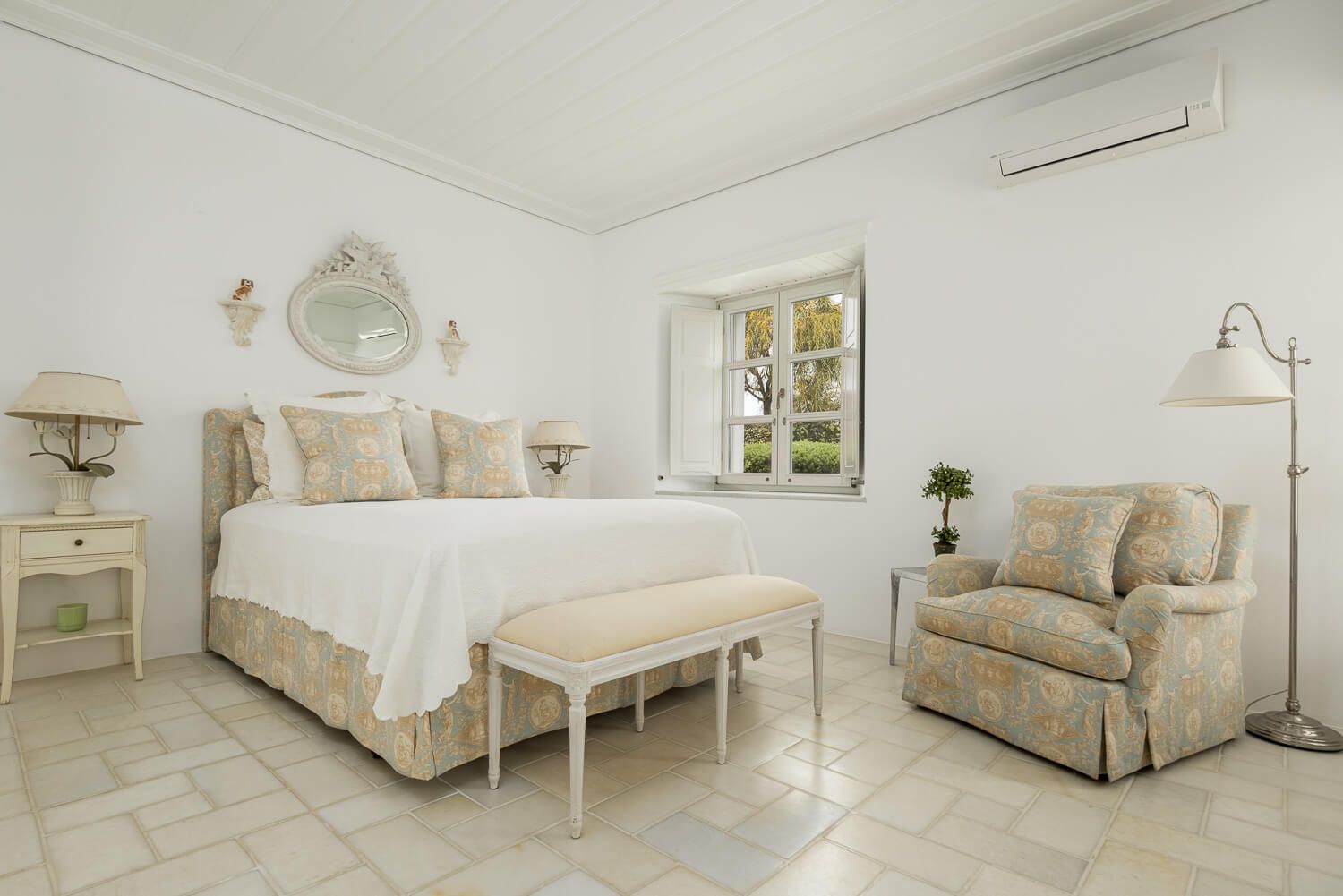 Elegant white bedroom with beige accents featuring a plush armchair, nightstand, and bench at the foot of the bed in Paros Greece