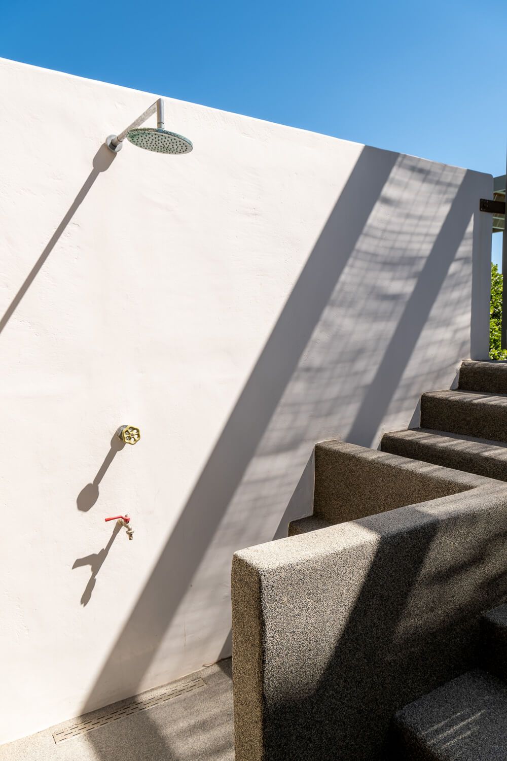 An outdoor shower with chrome fixtures mounted on a white wall next to concrete steps casting dramatic shadows.