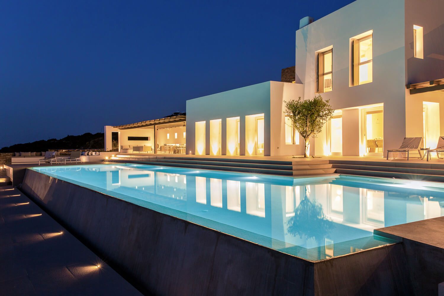Modern luxury villa with illuminated infinity pool overlooking ocean at twilight creates stunning reflections.