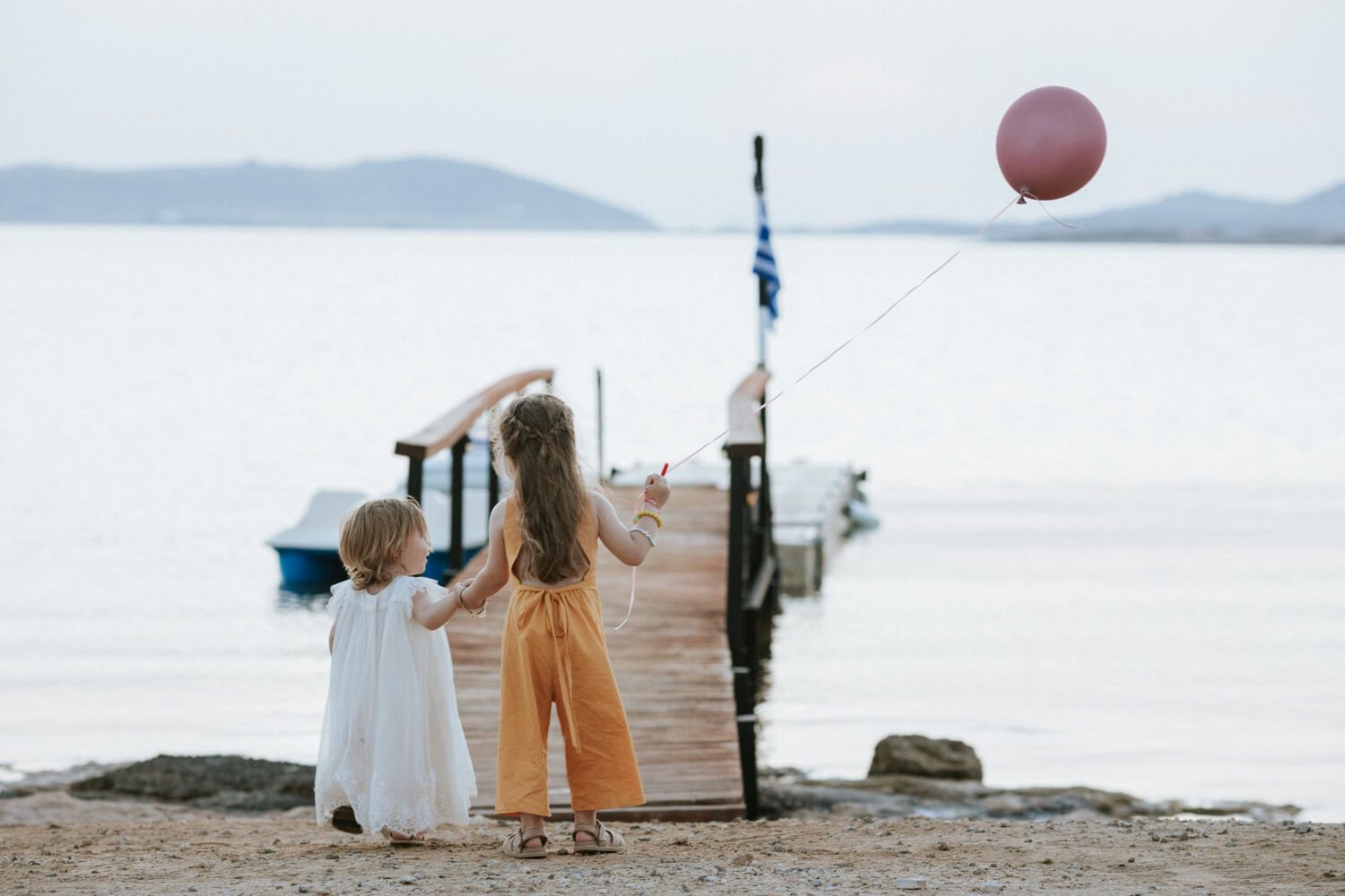 Two young children holding hands by a wooden pier, with a pink balloon floating above at Siparos.