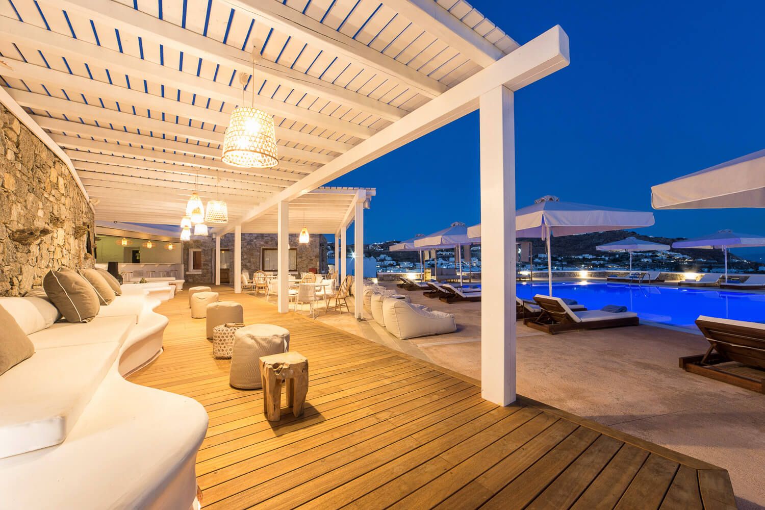 Luxurious outdoor terrace with white pergola, plush seating, and pool overlooking Mykonos bay at dusk.