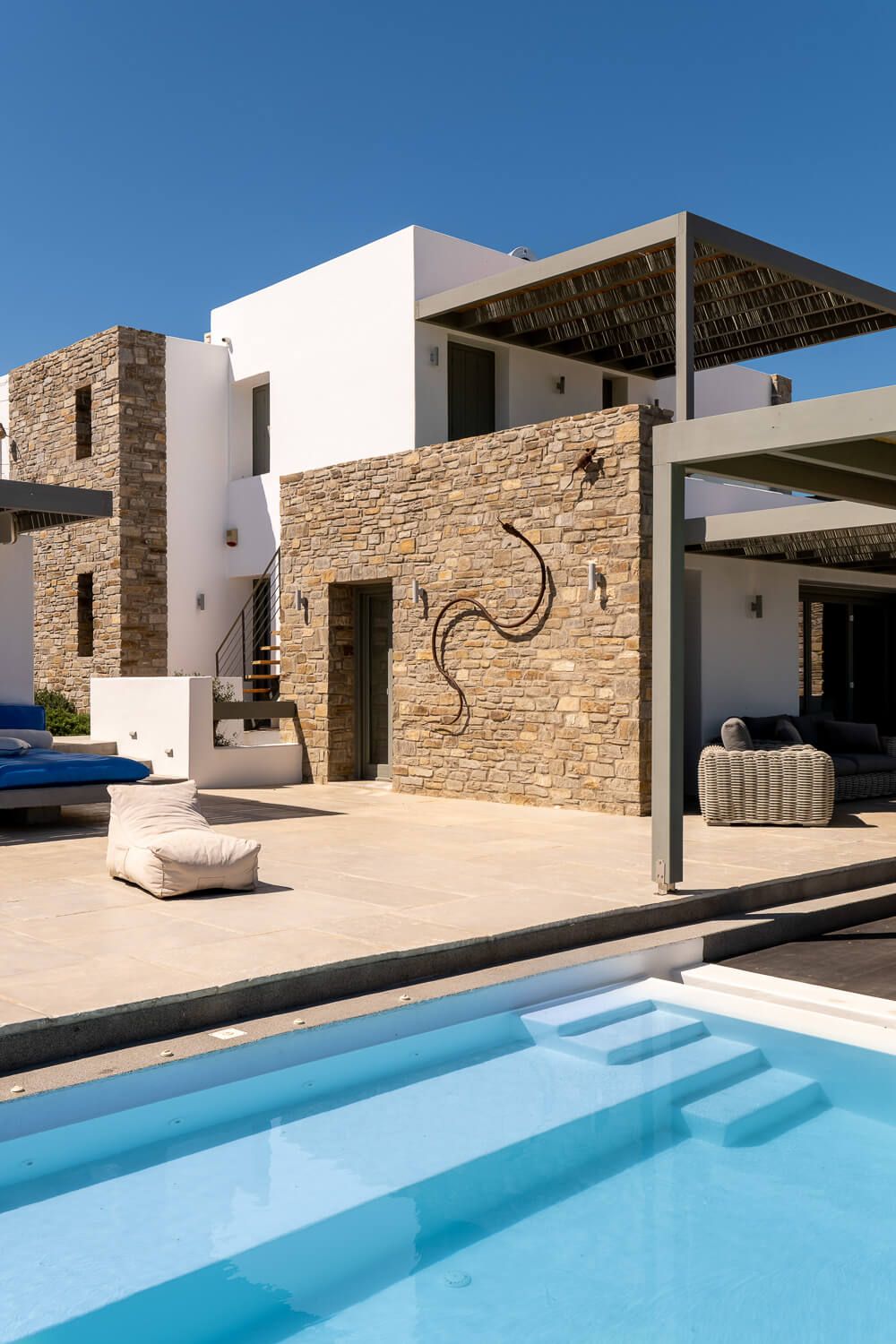 Modern luxury villa with stone walls and infinity pool overlooking blue water against white architecture in Mediterranean style.