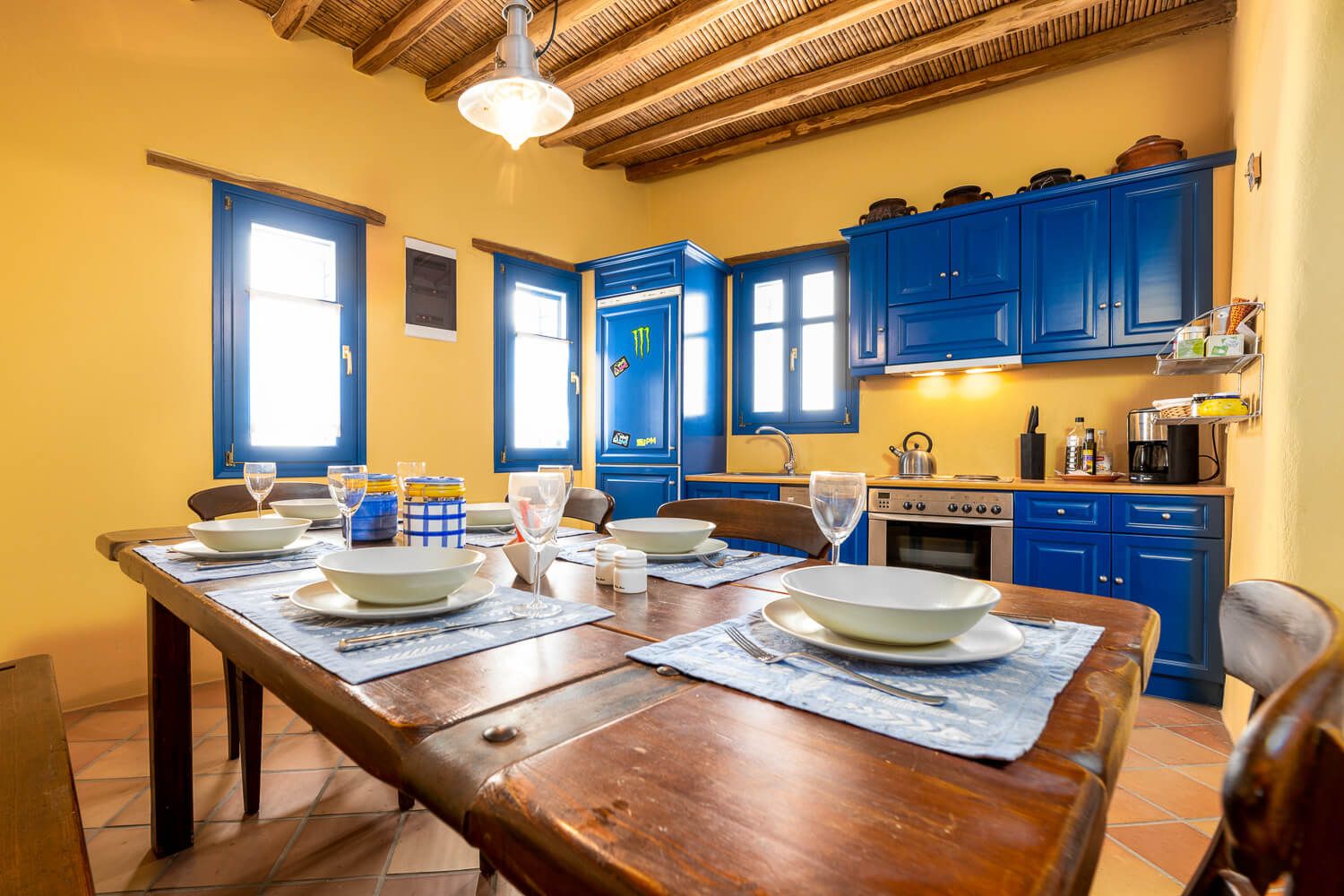 Rustic kitchen interior with exposed wooden beams, blue cabinets, and a long wooden dining table in warm lighting.