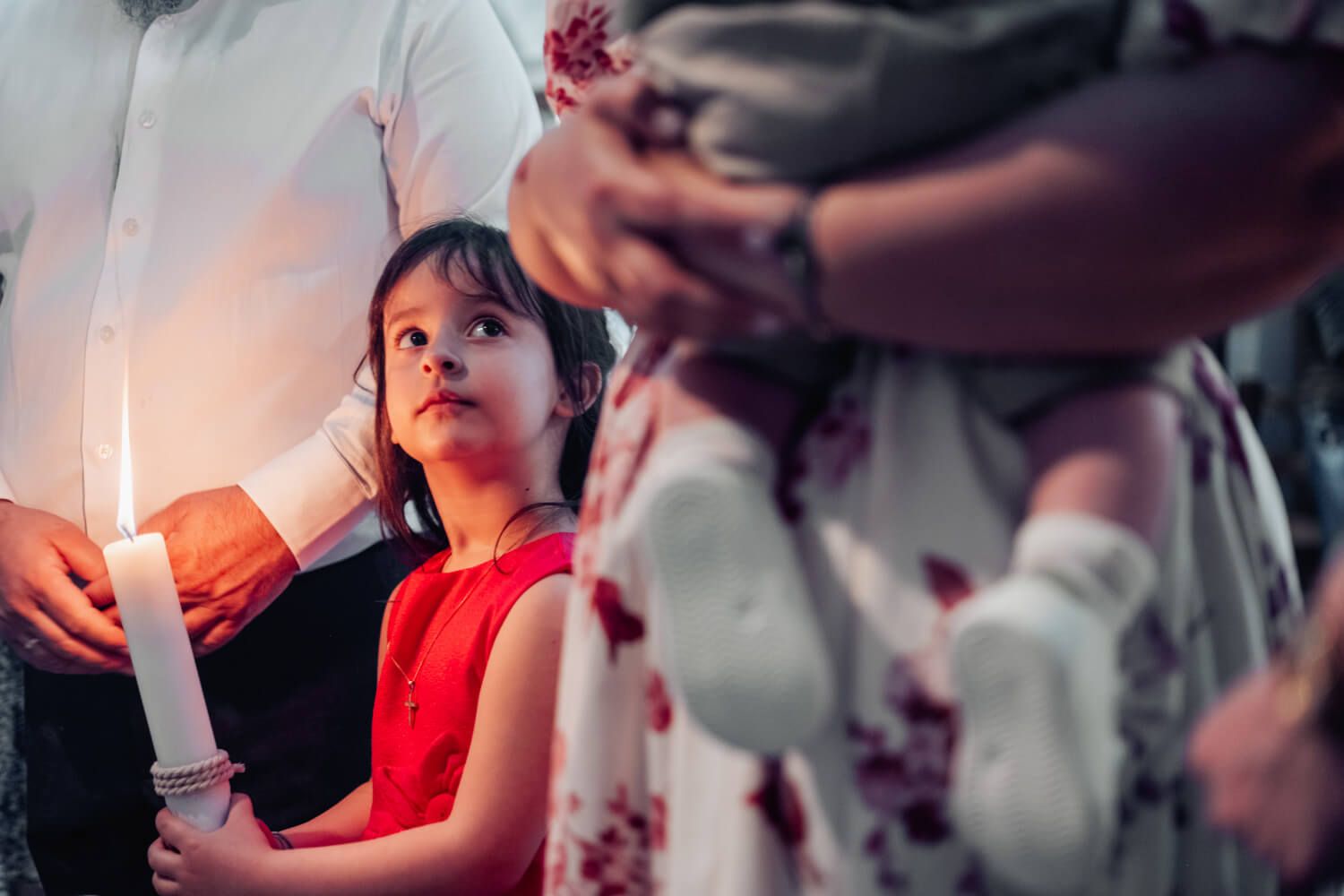 Young Asian girl in red dress holding white candle at religious ceremony with adults nearby.