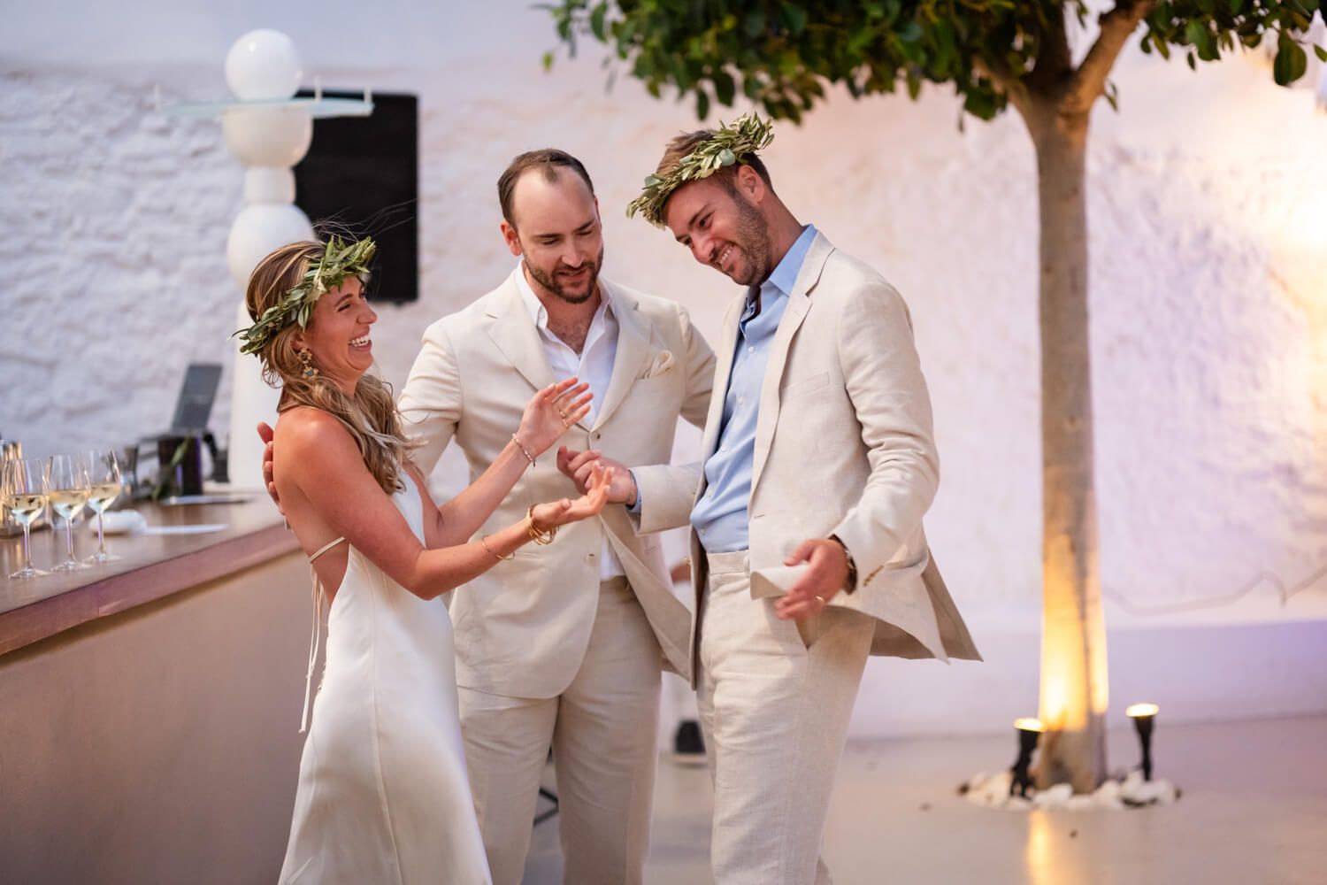 Bride and groom laugh together wearing floral crowns at outdoor Mediterranean wedding reception.