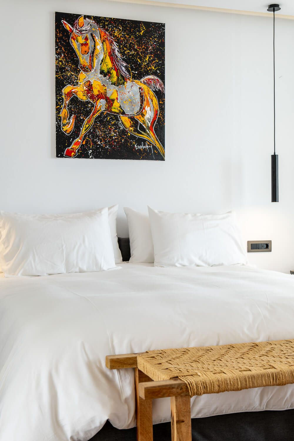 Modern villa room interior with white bedding, woven bench, and abstract orange and black artwork on the wall.