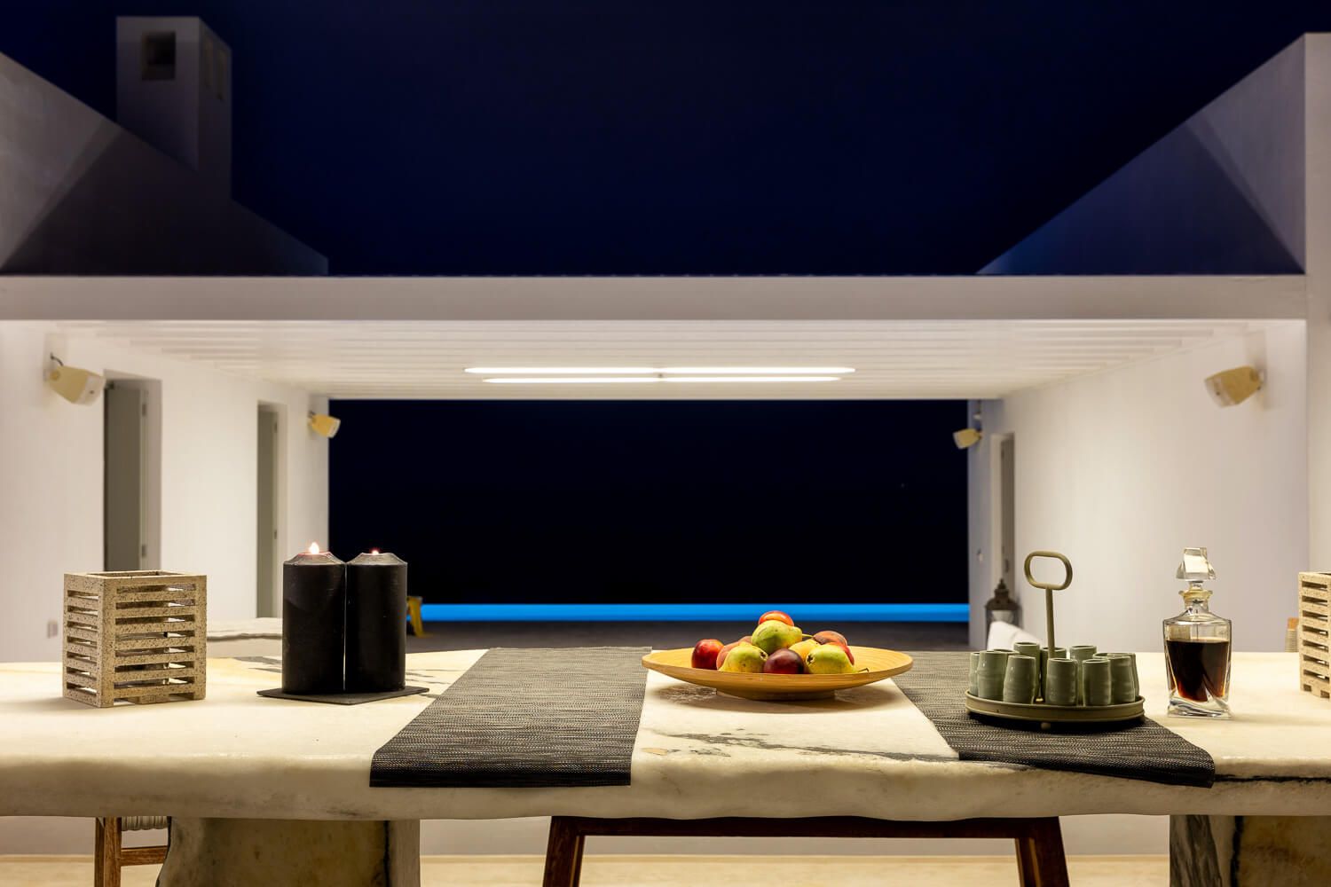 Elegant outdoor dining area with stone table, fruit bowl, and infinity pool overlooking night sky.