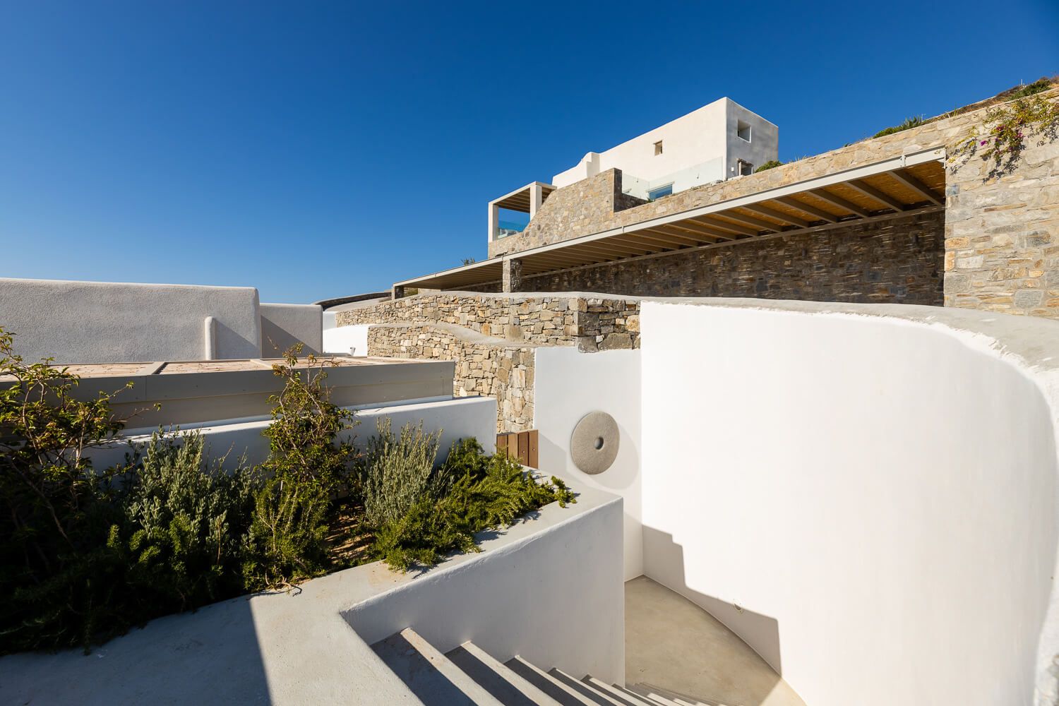 White Cycladic architecture with stone walls and curved staircases under a clear blue sky.