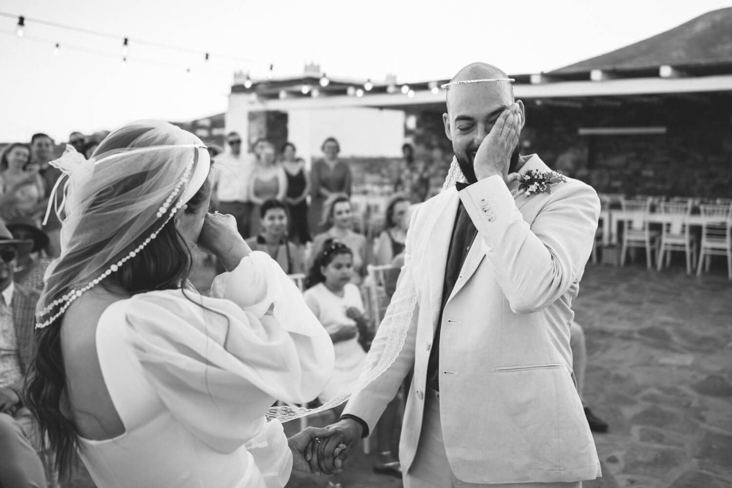 Black and white photo of emotional groom wiping tears during heartfelt outdoor wedding ceremony with guests watching.