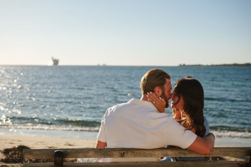 Romantic couple sitting close together on a bench facing the calm blue sea at sunset.