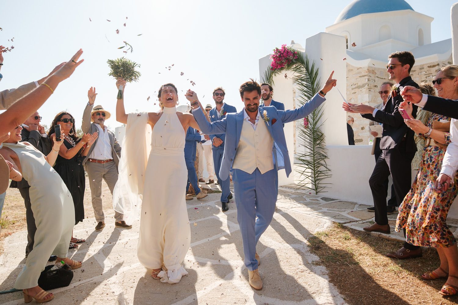 Joyful wedding exit at Agios Fokas Paros - Couple walking through olive leaves towards the boat