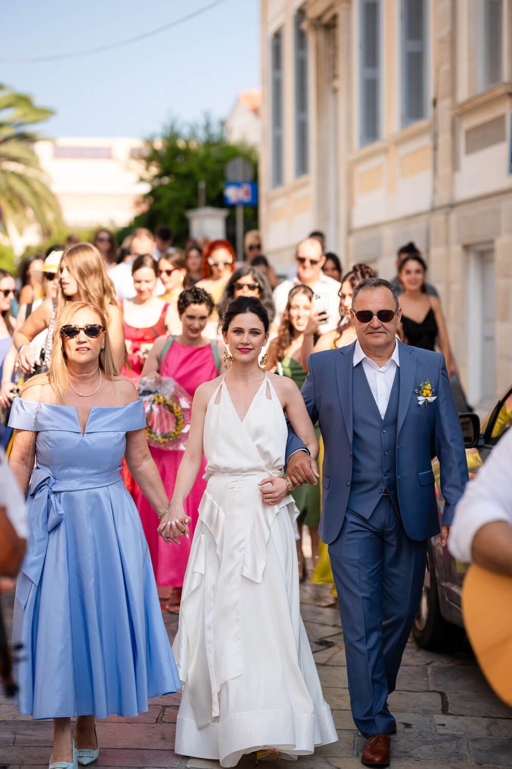 Wedding party walks down sunny street in Syros Greece with mother and father and bride in white gown