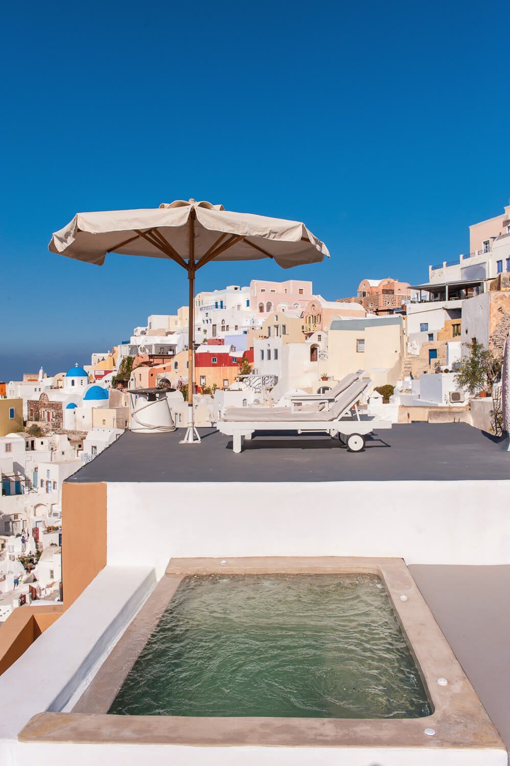 Luxurious infinity pool with beige umbrella overlooking white Santorini buildings and blue Mediterranean Sea on sunny day.