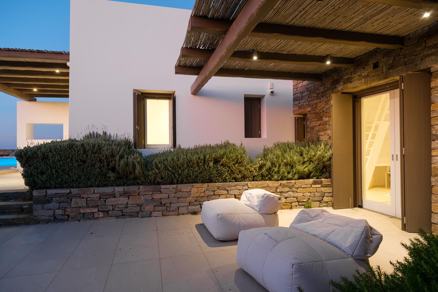Modern luxury villa with multiple outdoor seating areas and white cushioned furniture on a stone patio at sunset.