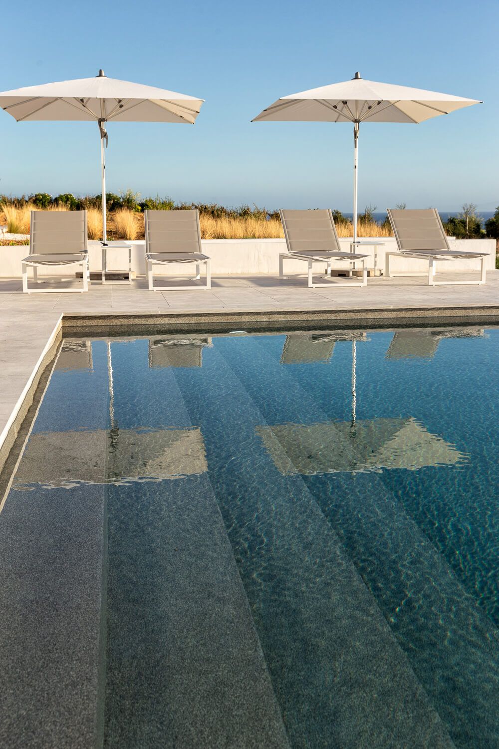 Poolside with white lounge chairs and umbrellas reflecting in blue water under clear sky in Mykonos.