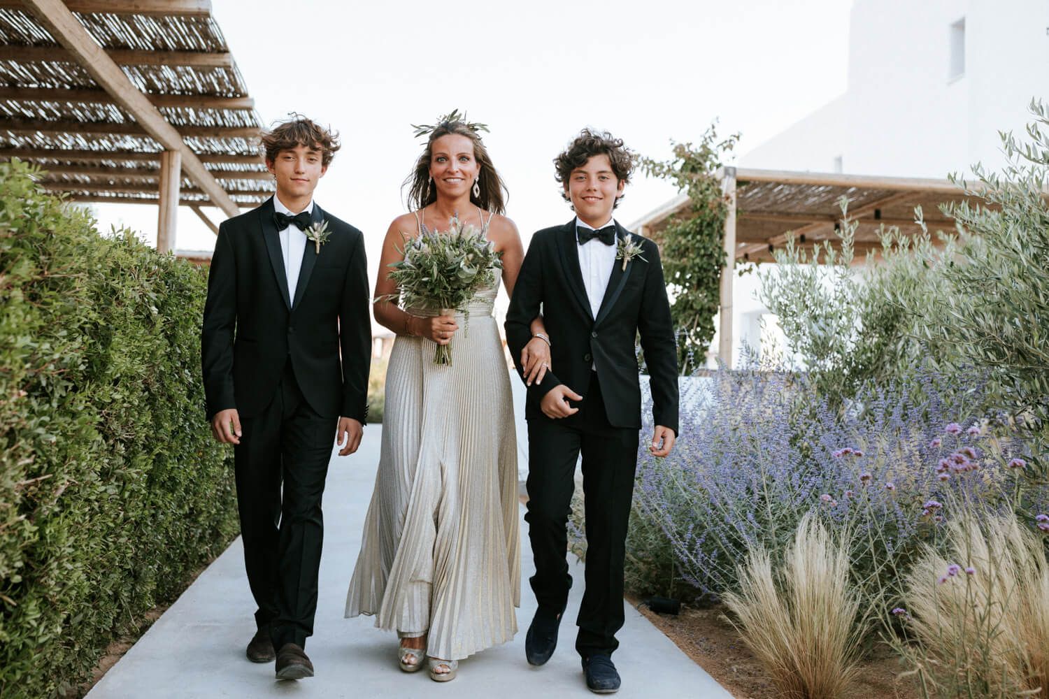 A bride in a flowing gown walks with two young men in tuxedos through a sunny outdoor wedding venue.