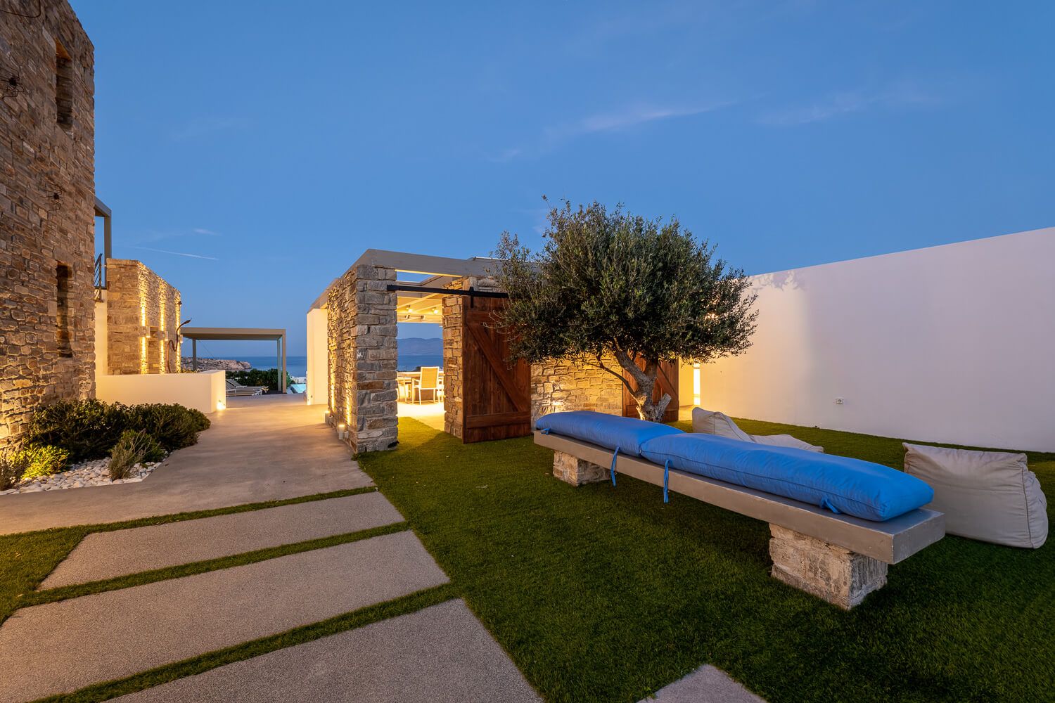 Modern outdoor patio with illuminated stone pathway, blue cushioned bench and olive tree at dusk.
