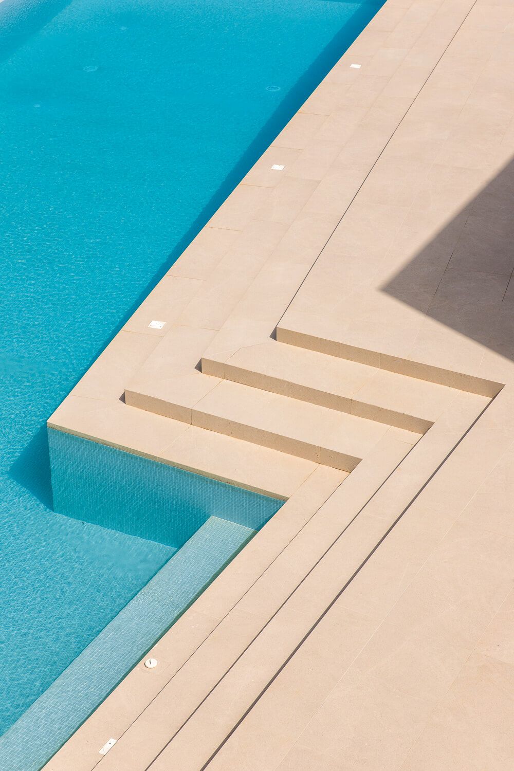 Modern swimming pool design featuring geometric steps and turquoise water against beige concrete decking in minimalist style.