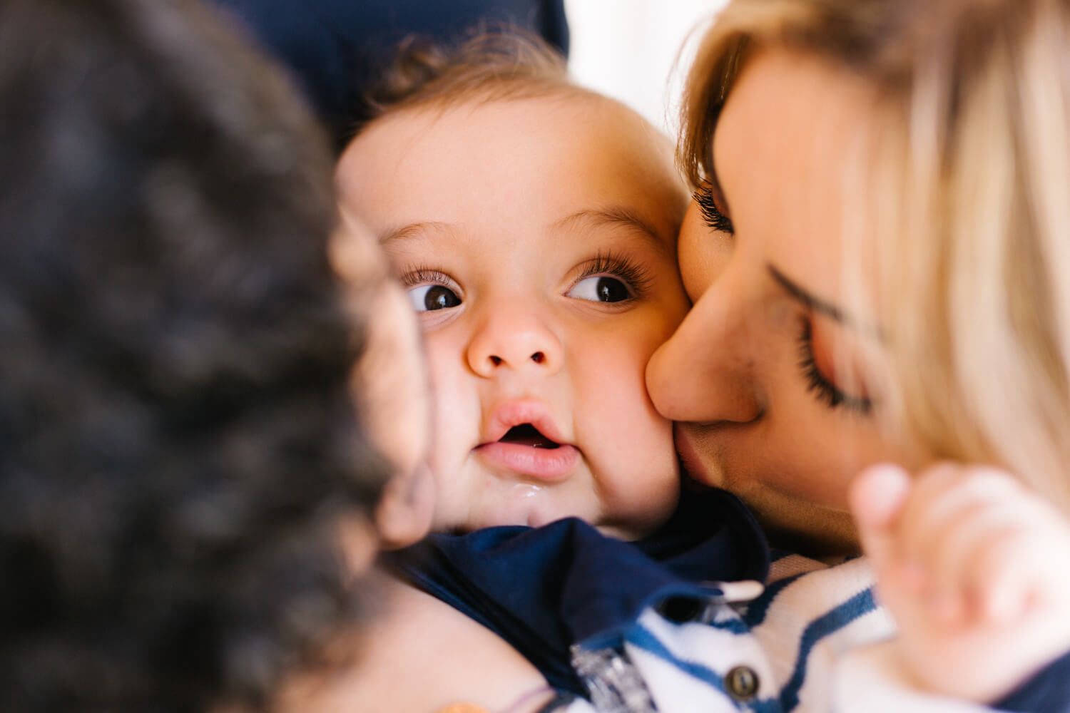 Parents tenderly kiss a wide-eyed baby captured in a warm, intimate close-up photo.