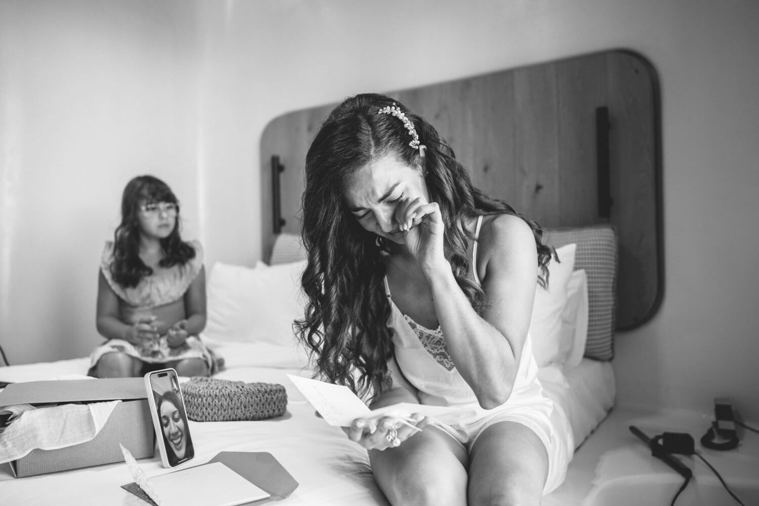 Emotional bride reading a letter on a bed while a bridesmaid sits in the background, black and white