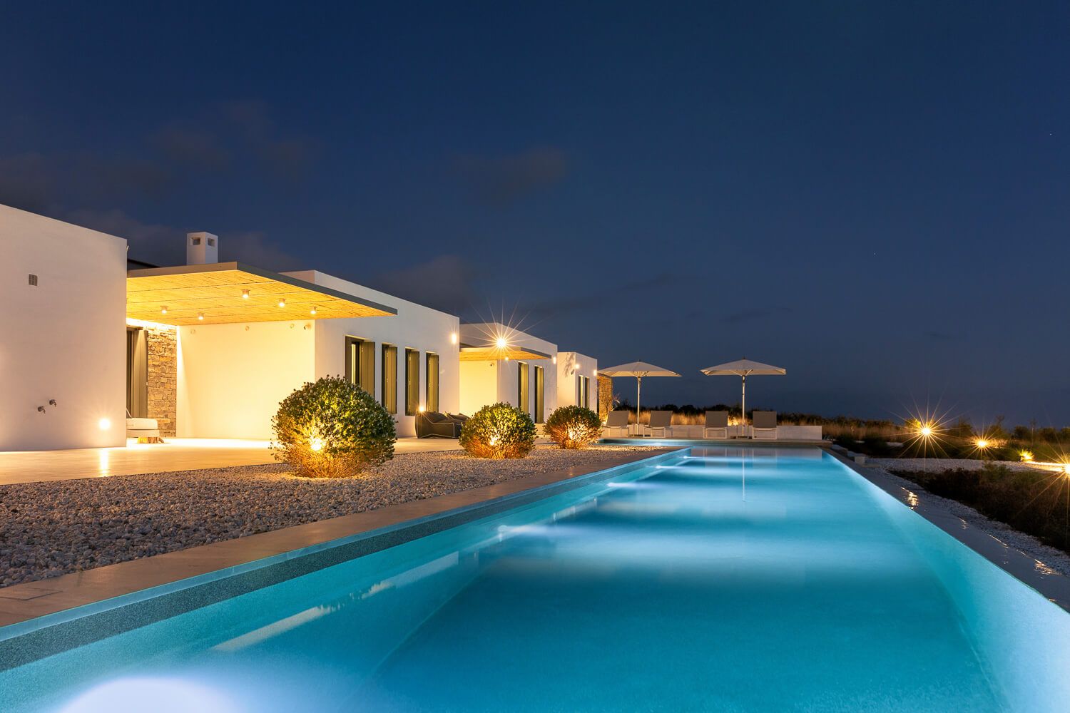 Luxury villa with illuminated pool at night, white architecture, and landscaped gardens in Paros.