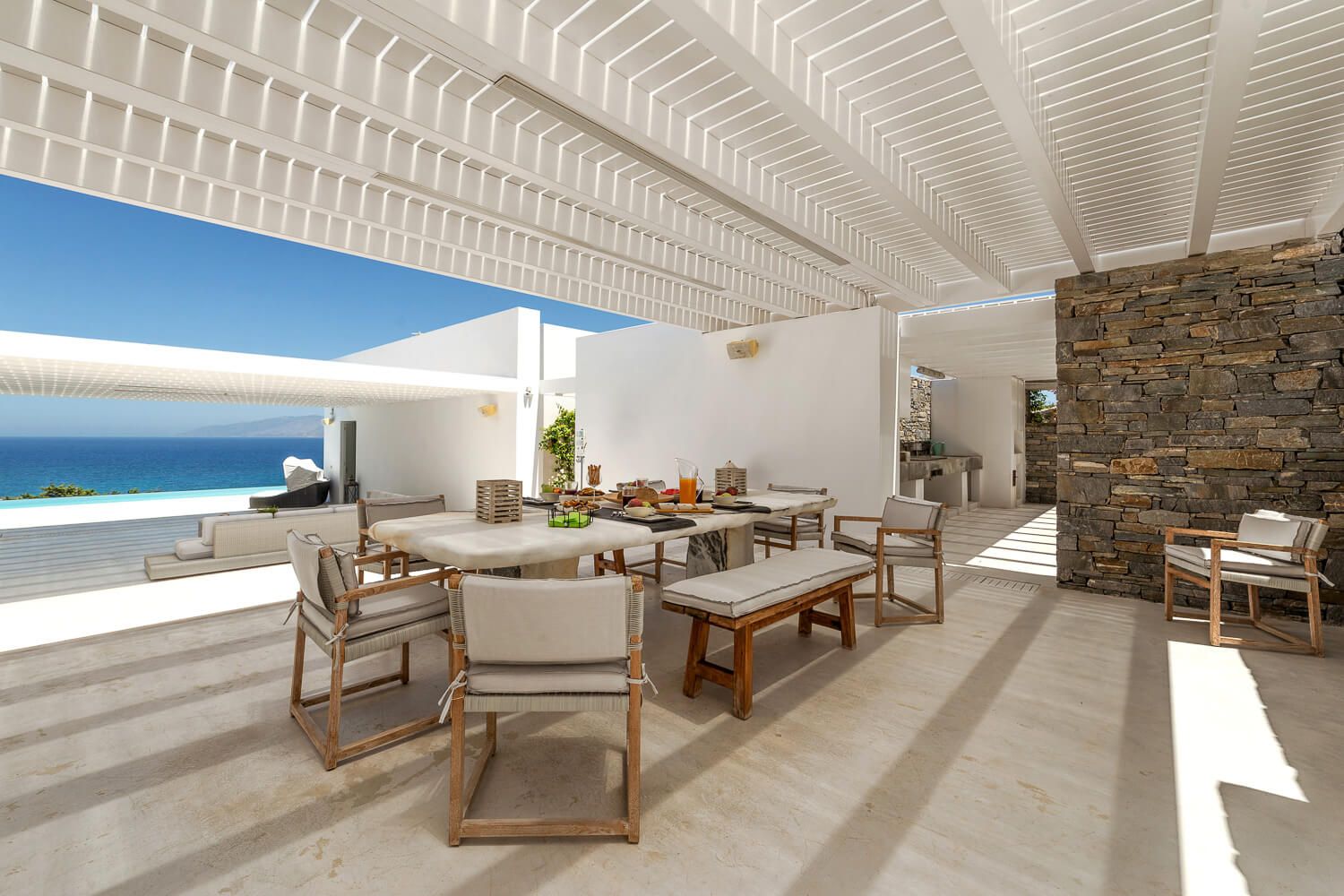 Luxurious outdoor terrace with teak furniture, pergola ceiling, stone wall, and stunning ocean views