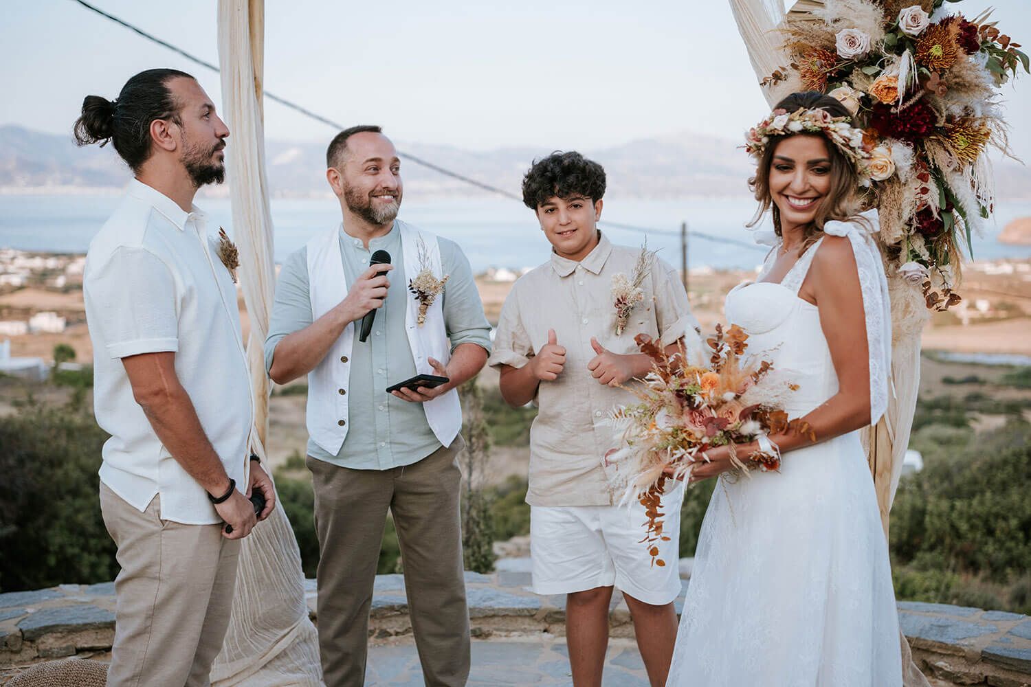 A wedding party laughs together on a sunny day wearing white and beige attire with autumn-colored floral bouquets.