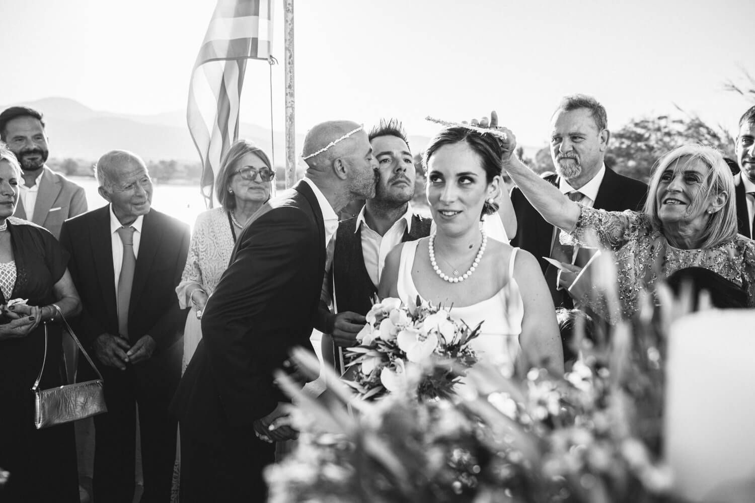 Black and white wedding photo of bride receiving a kiss from guest outdoors, holding bouquet, surrounded by smiling attendees.