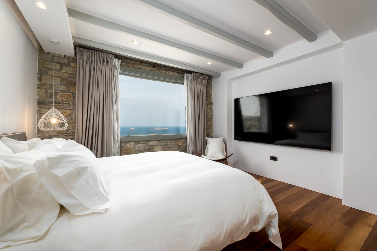 Modern bedroom with white bedding, hardwood floors, wall-mounted TV, and large window offering scenic ocean views