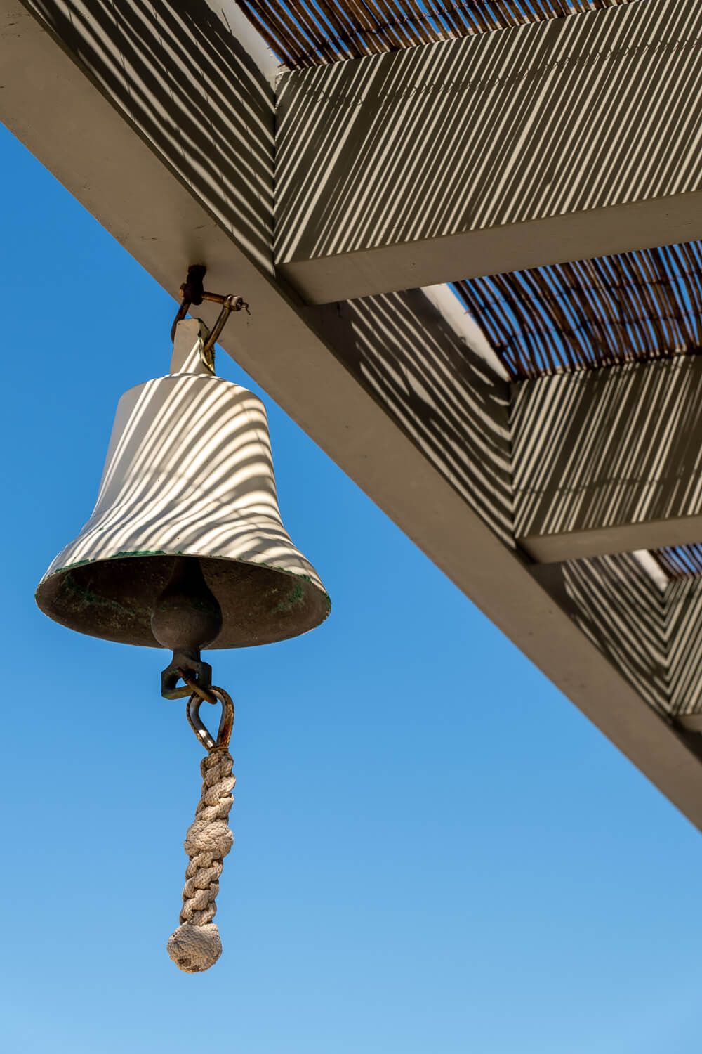 A white nautical bell with rope pull hangs against a bright blue sky beneath a slatted wooden overhang.