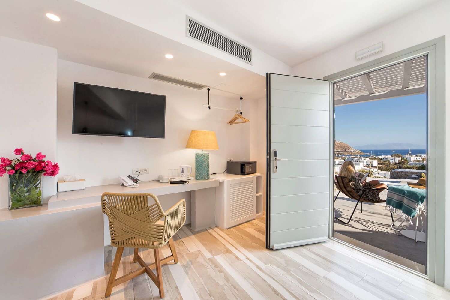 Modern hotel room with desk area and balcony overlooking ocean view, featuring light wood floors and contemporary decor.
