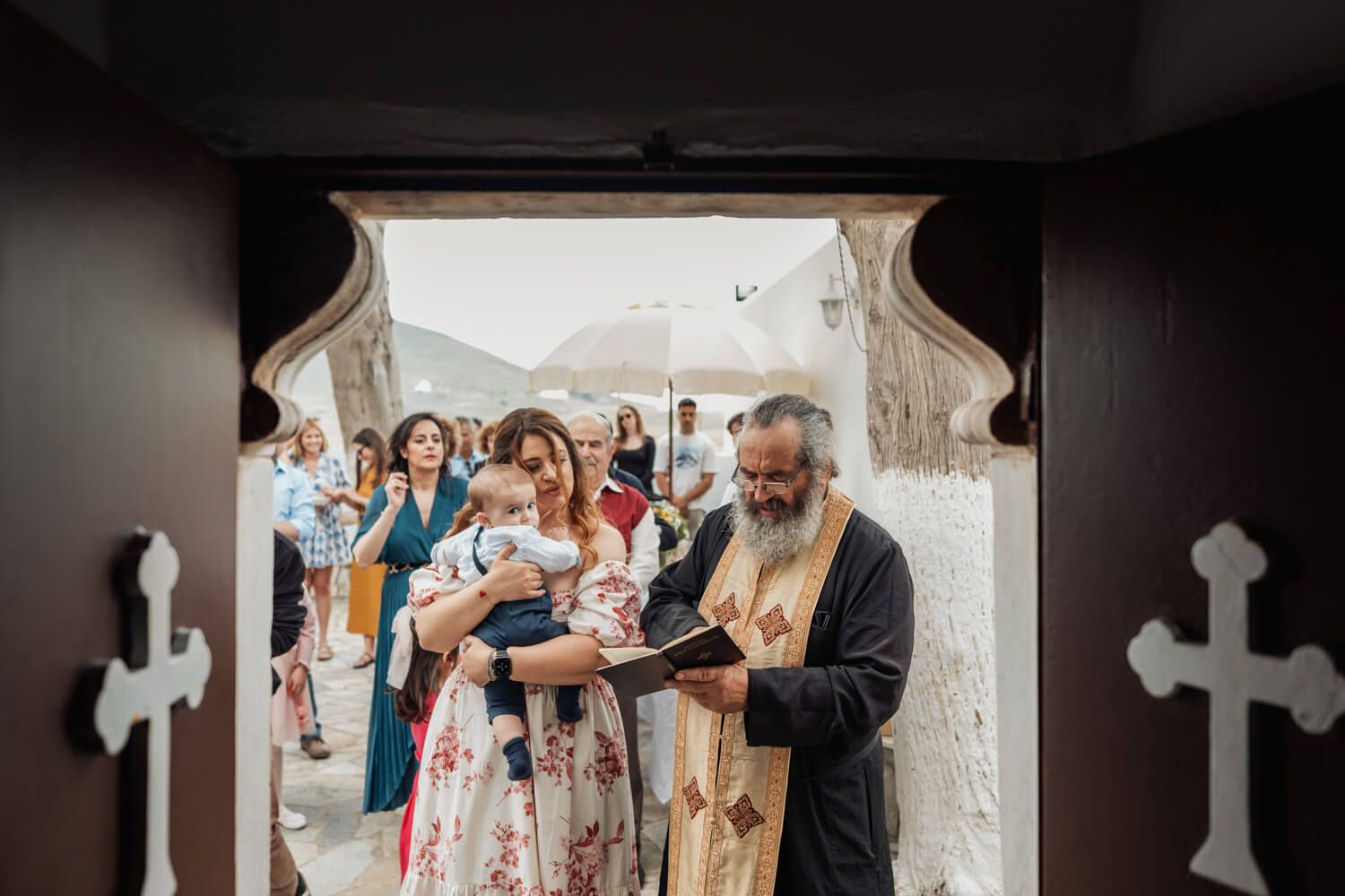 Priest reads scripture at church entrance during a baptism ceremony with mother holding infant.
