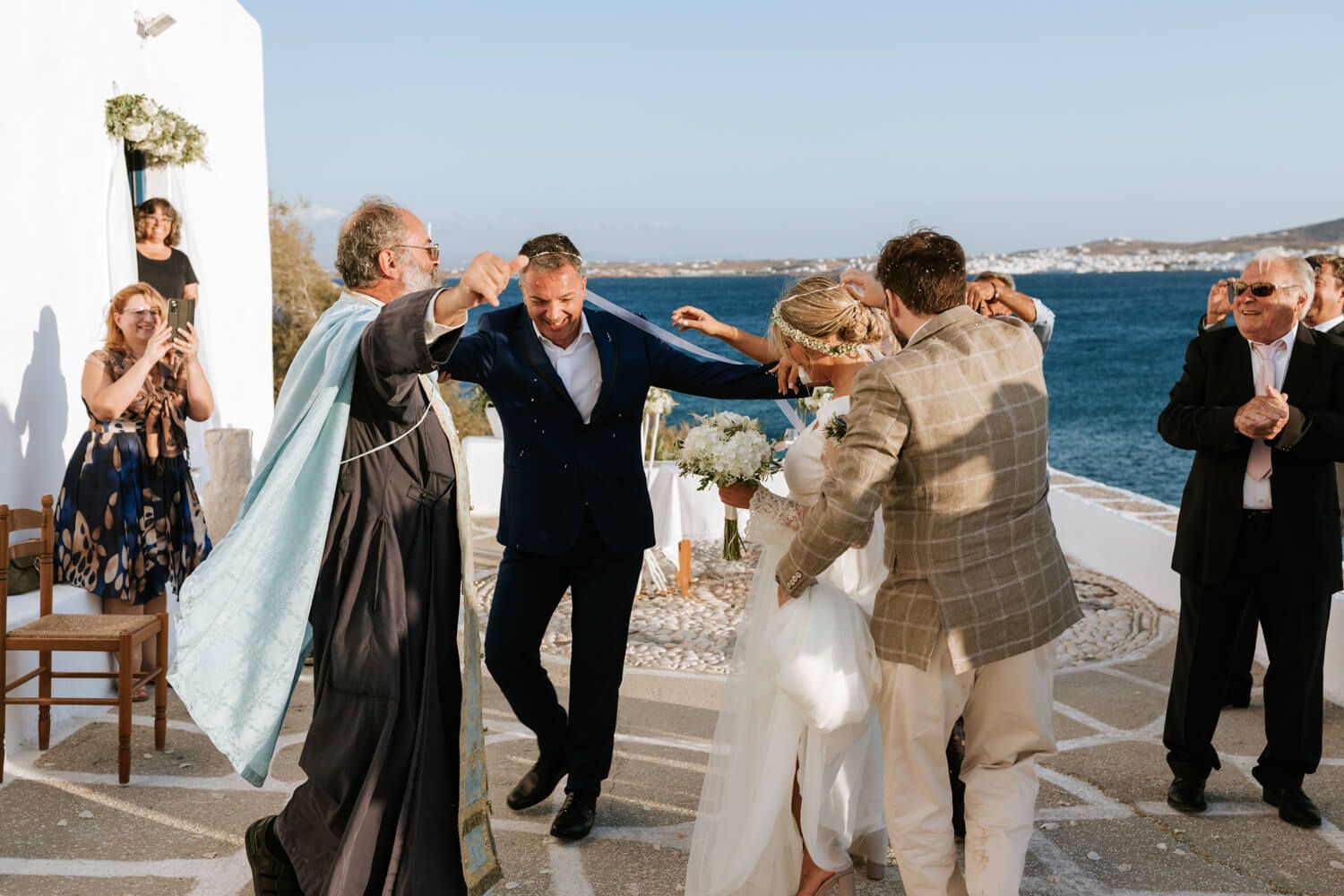 Outdoor Greek wedding ceremony by the sea with priest, bride, groom, and guests celebrating in Mykonos.