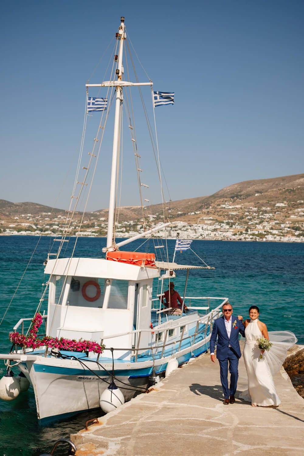 Bride and groom walking on a dock beside a Greek fishing boat decorated with flowers and flags.
