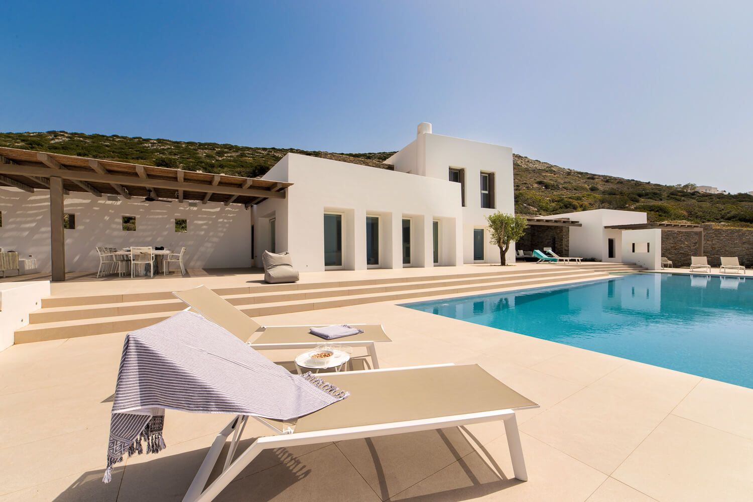Luxurious white Mediterranean villa with modern sun loungers beside a sleek swimming pool against hillside backdrop.