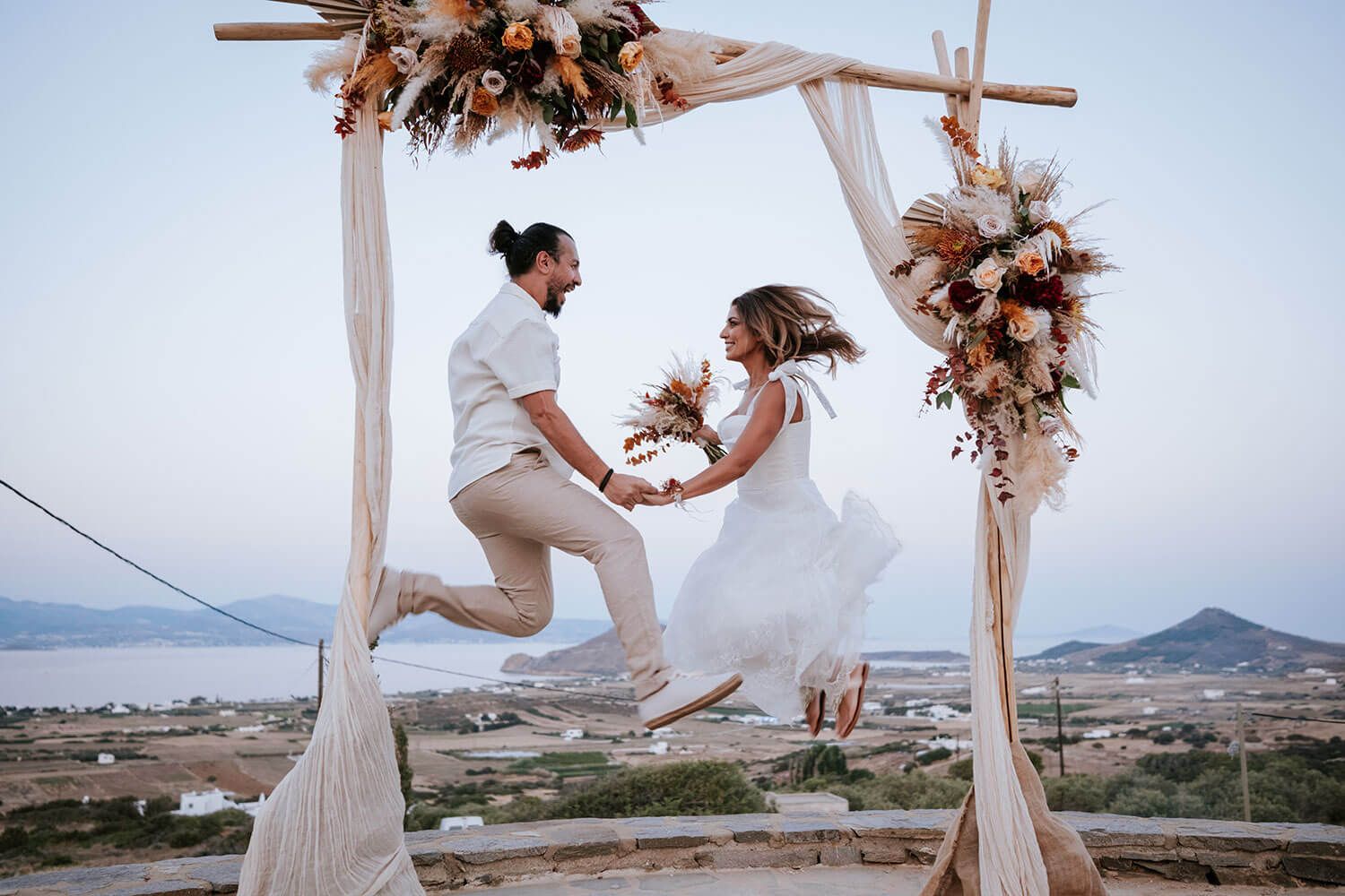 The newlyweds jump in the air under a romantic outdoor wedding ceremony setup with flowing white fabric and dried floral arrangements